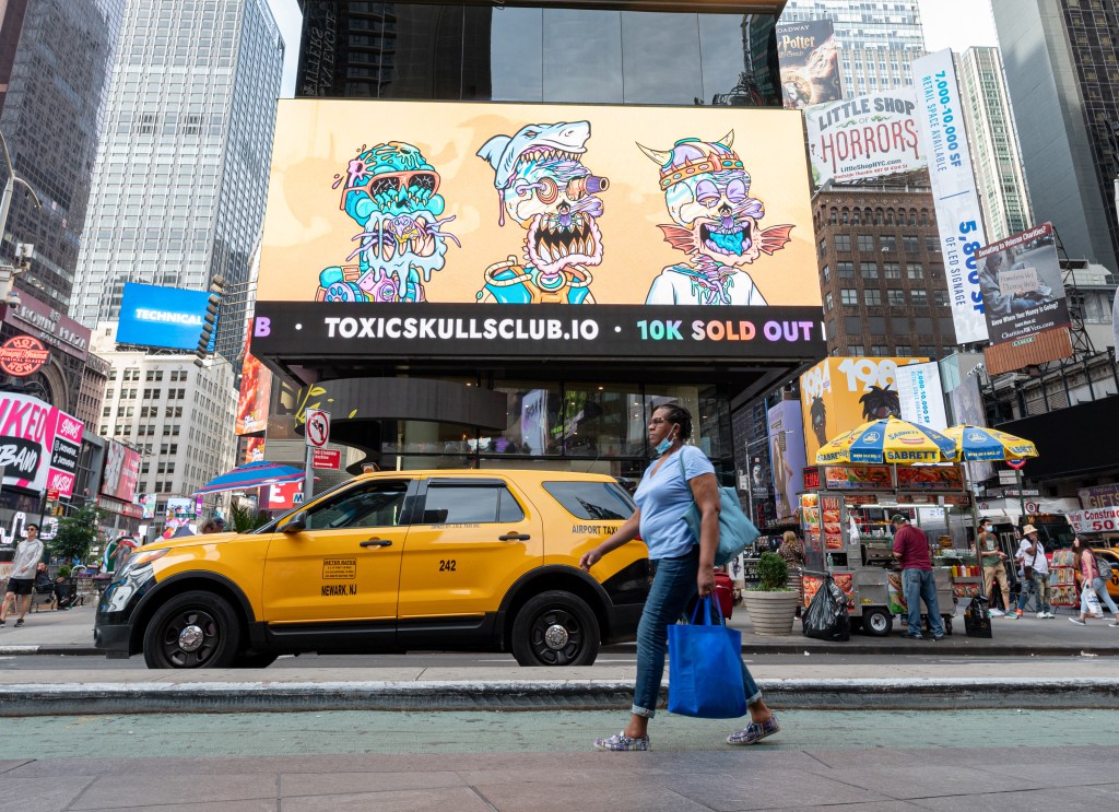 A person walks by a "Toxic Skulls Club" NFT billboard in Times Square while a taxi drives by.